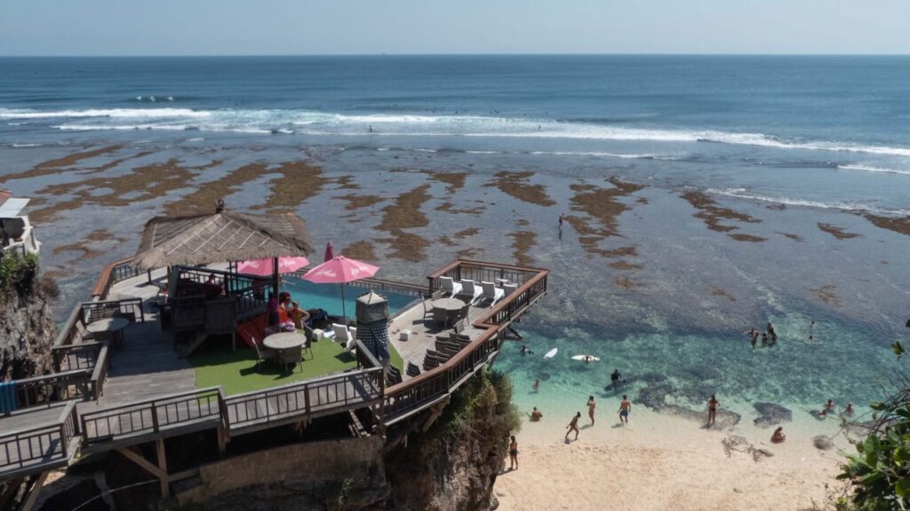 A pool and lounge platform built above a Balinese beach. On the beach people are paddling in the lagoon formed by an outgoing tide. Surfers are in the distance.